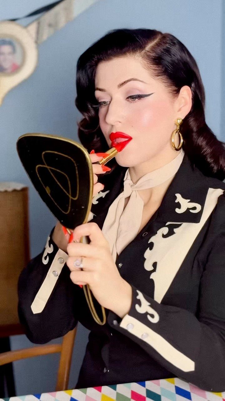 1950s Day Dress Ideas: 6 Retro Looks, From Gingham to Florals 1 Woman with vintage hair applying bright red lipstick using a handheld mirror