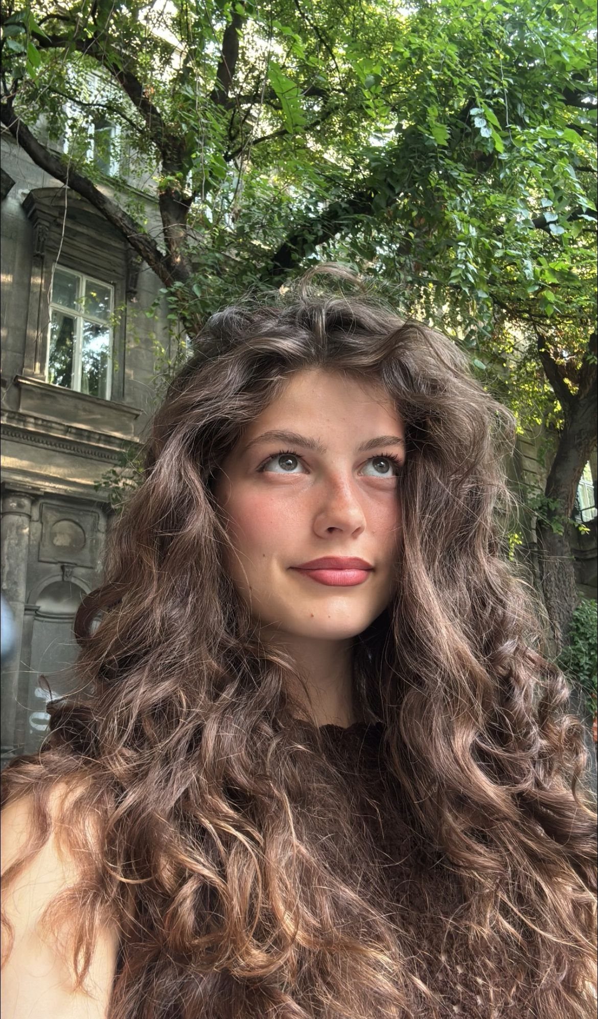 Woman with long, voluminous wavy brown hair posing against a backdrop of trees.