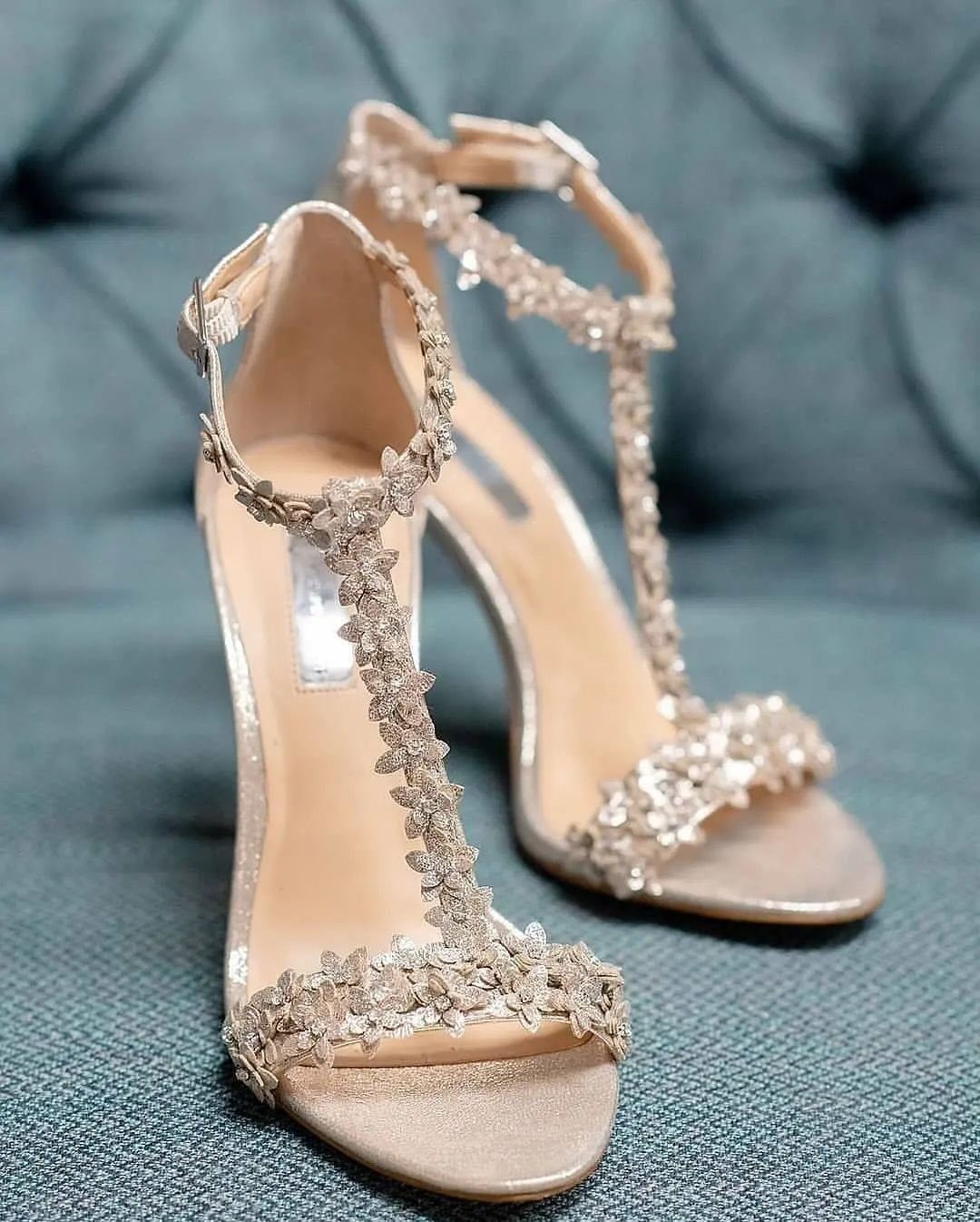 Nude high-heeled sandals with floral embellishments and ankle straps on grey fabric.