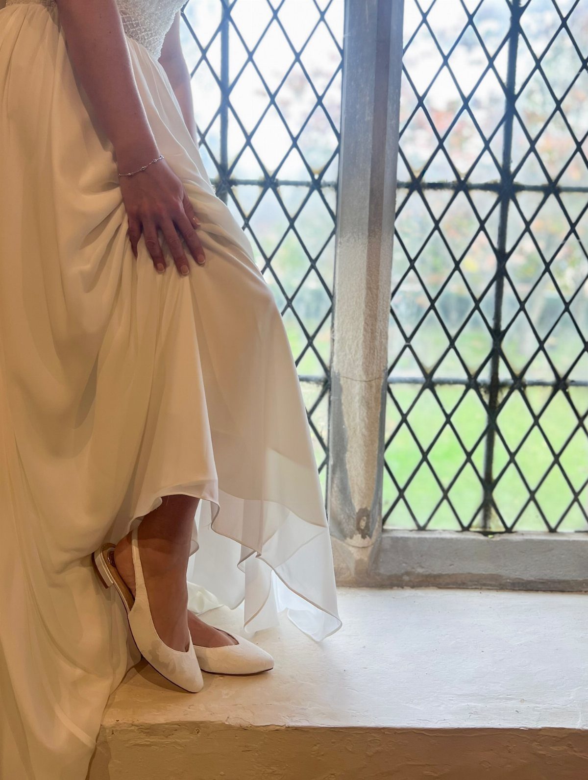 White pointed-toe wedding pumps worn with a flowing white bridal gown by a window