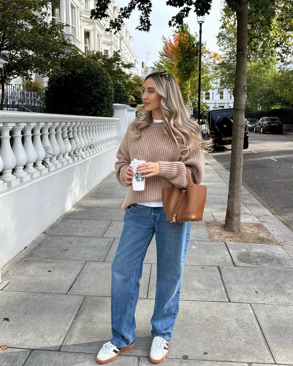 Woman in beige oversized sweater, wide-leg blue jeans, and white sneakers on sidewalk