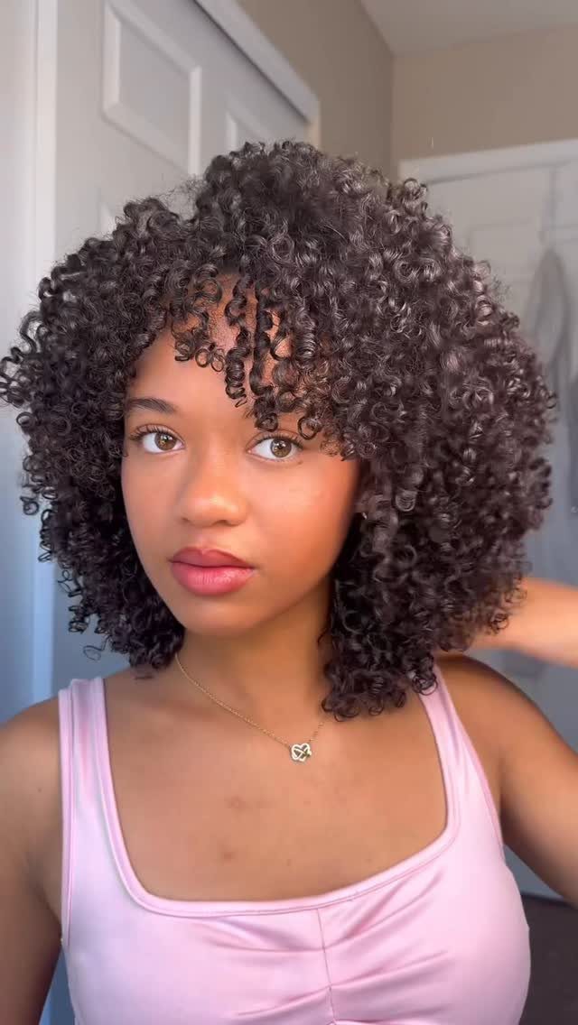 Woman with voluminous tight black curly hair and bangs wearing a pink top.