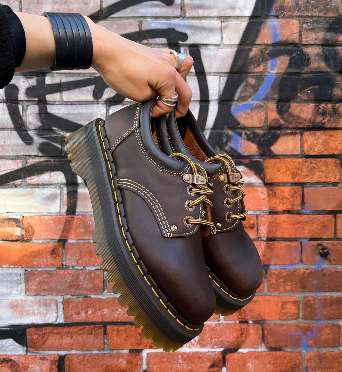 Brown leather platform casual shoes with yellow stitching held against a brick wall