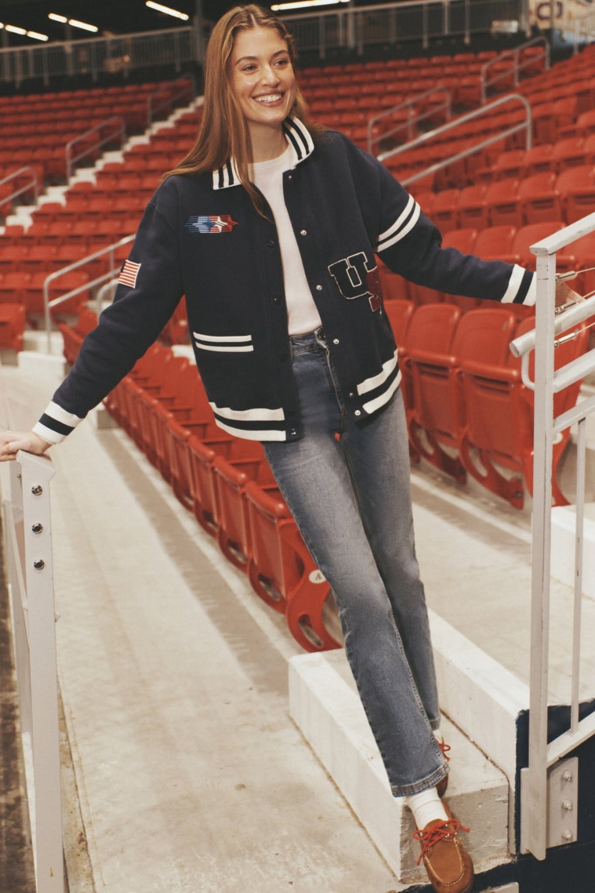 Woman wearing a navy and white varsity jacket with blue jeans in a stadium