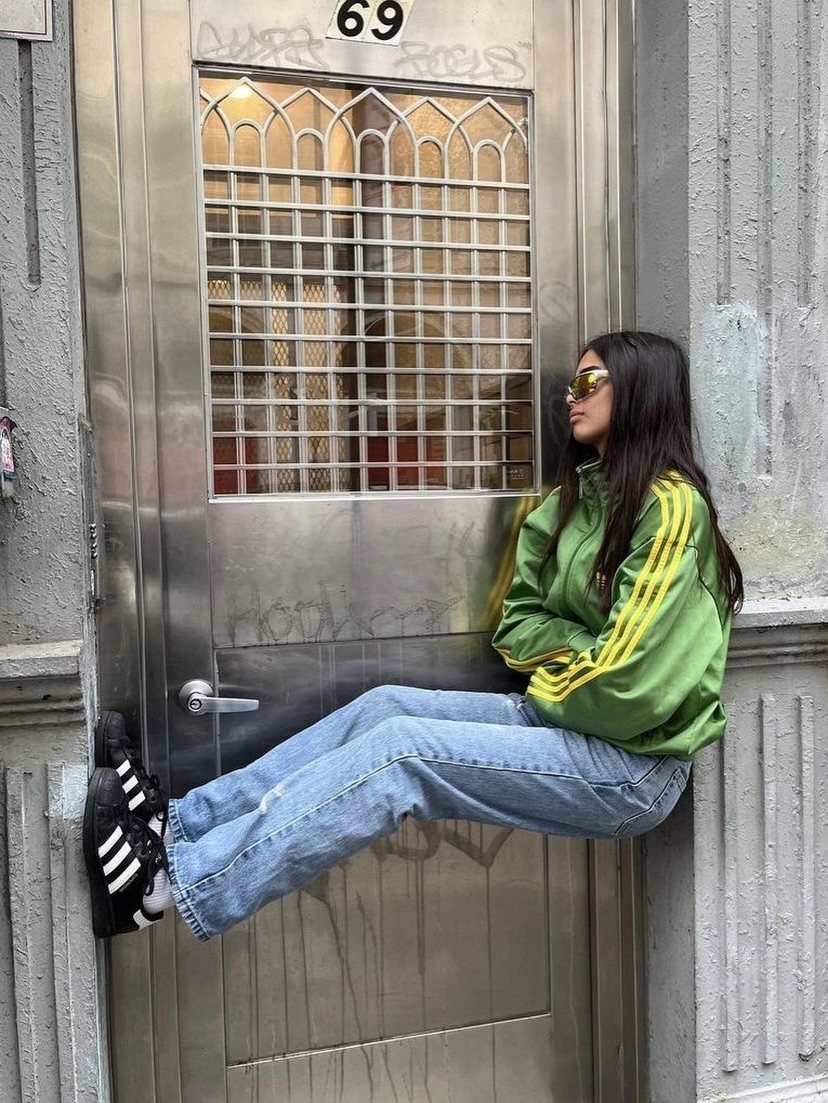 Woman wearing green Adidas track jacket and blue jeans sitting by a door