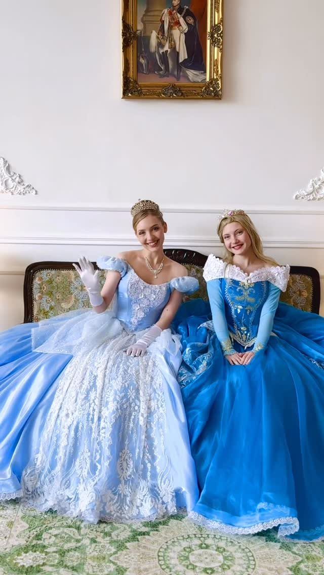 Two women wearing blue Cinderella-style ball gowns and tiaras sitting on a sofa.