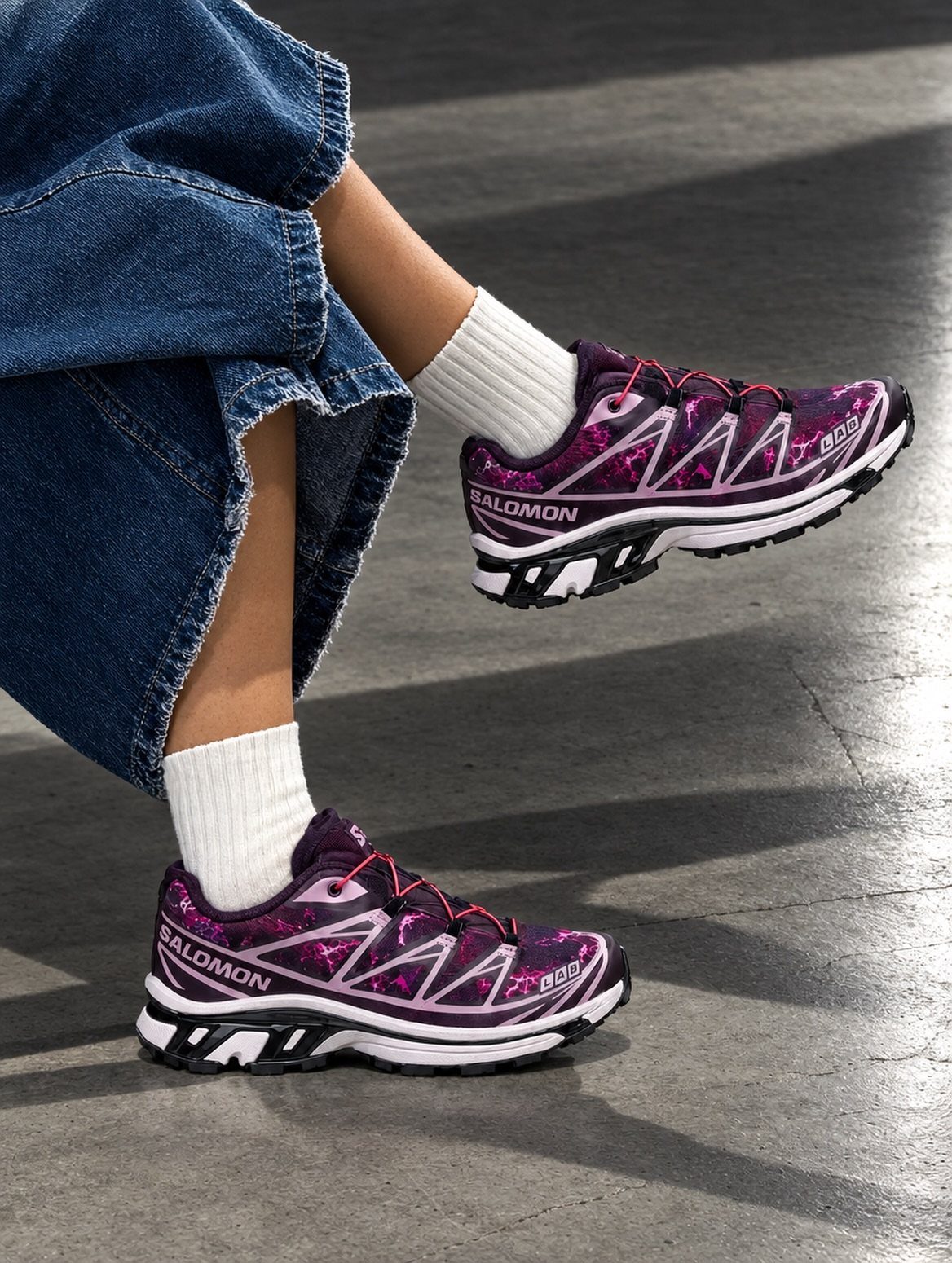 Purple patterned Salomon sneakers with white socks and blue denim jeans on concrete.