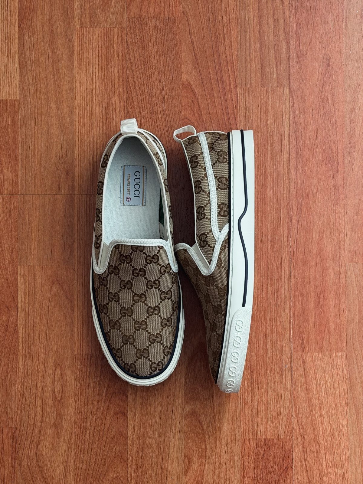 Brown monogrammed slip-on sneakers with white soles on a wooden floor.