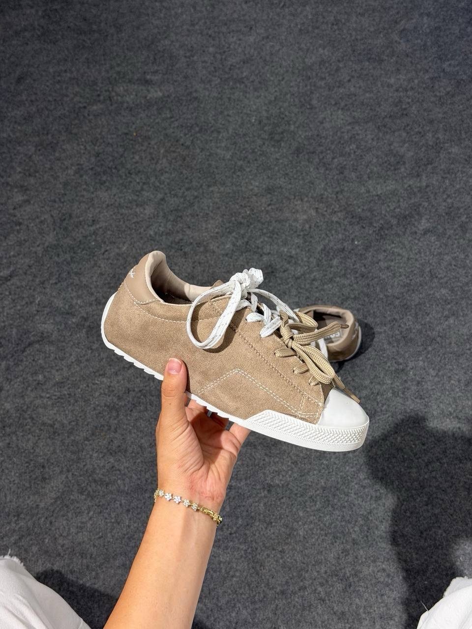 Tan suede sneaker with white laces held by a hand over grey pavement.