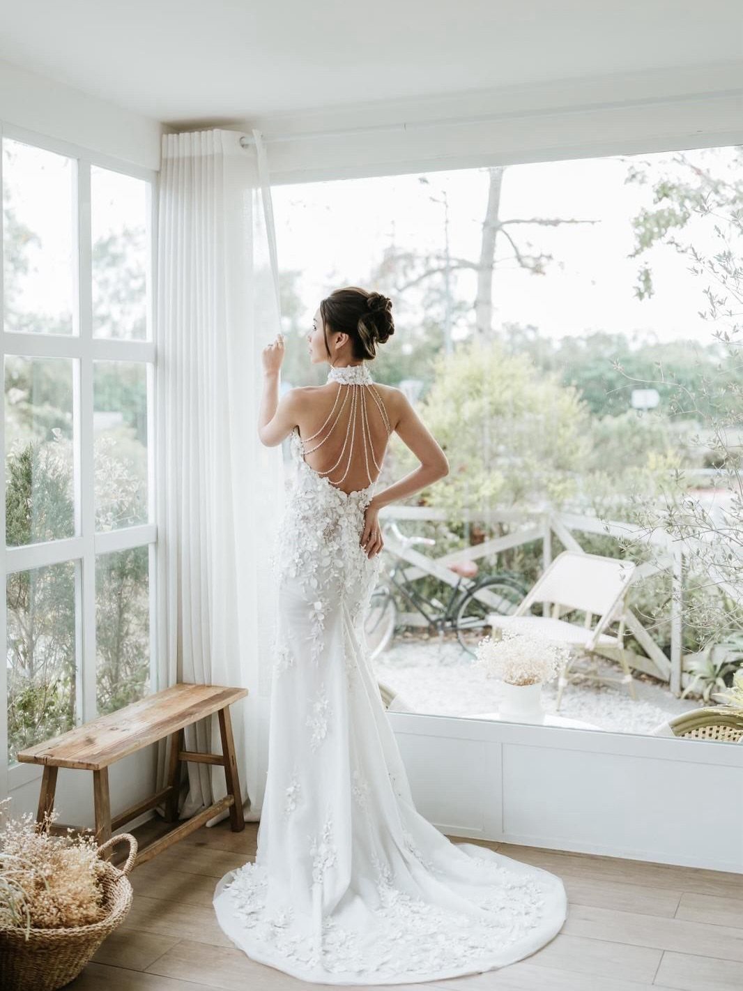 Woman in a white lace halter neck backless wedding dress standing by a window