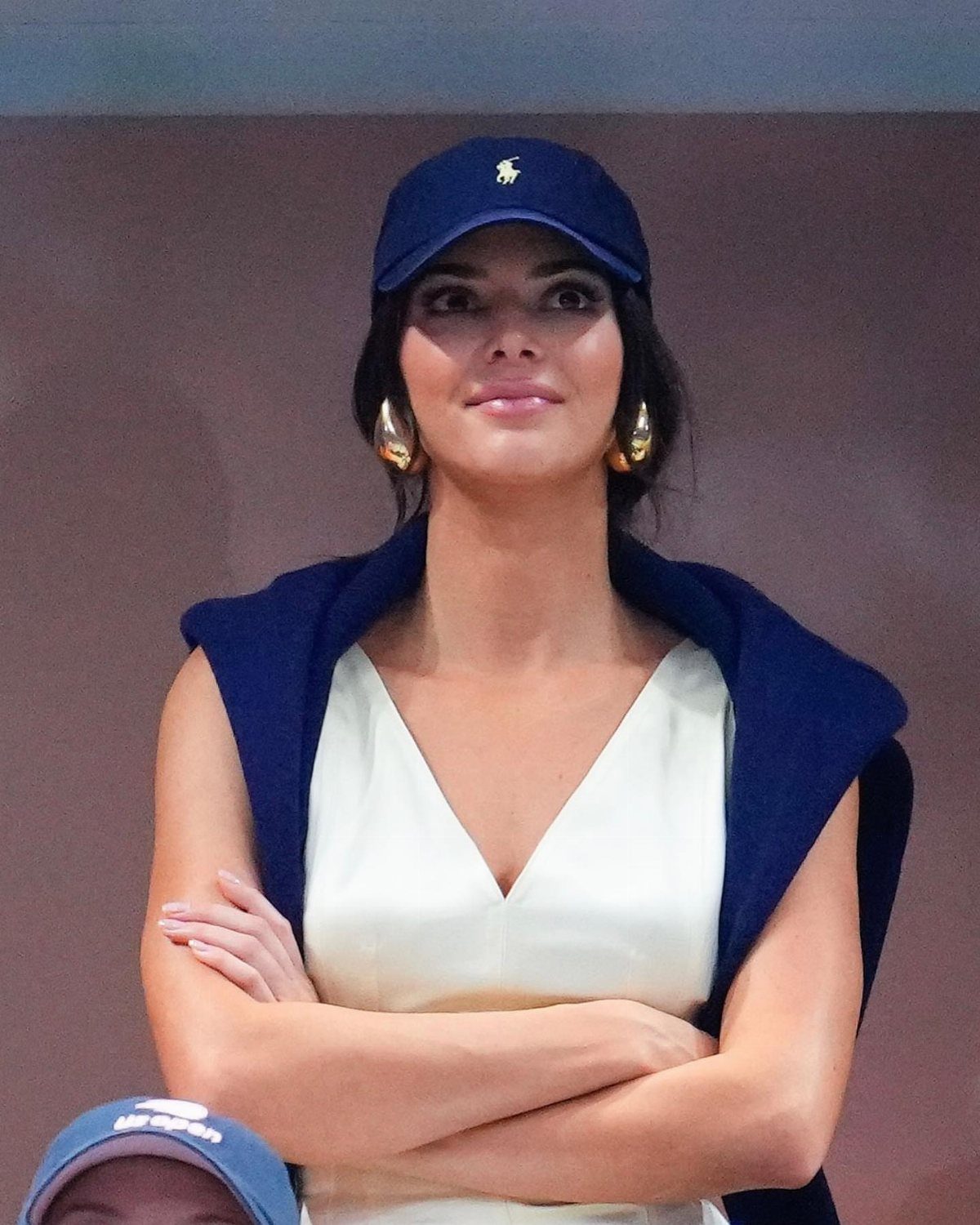 Woman wearing a blue baseball cap, white top, and blue sweater over shoulders.