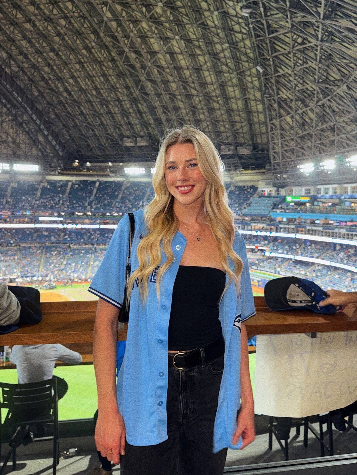 Blonde woman wearing a light blue baseball jersey and black jeans at stadium