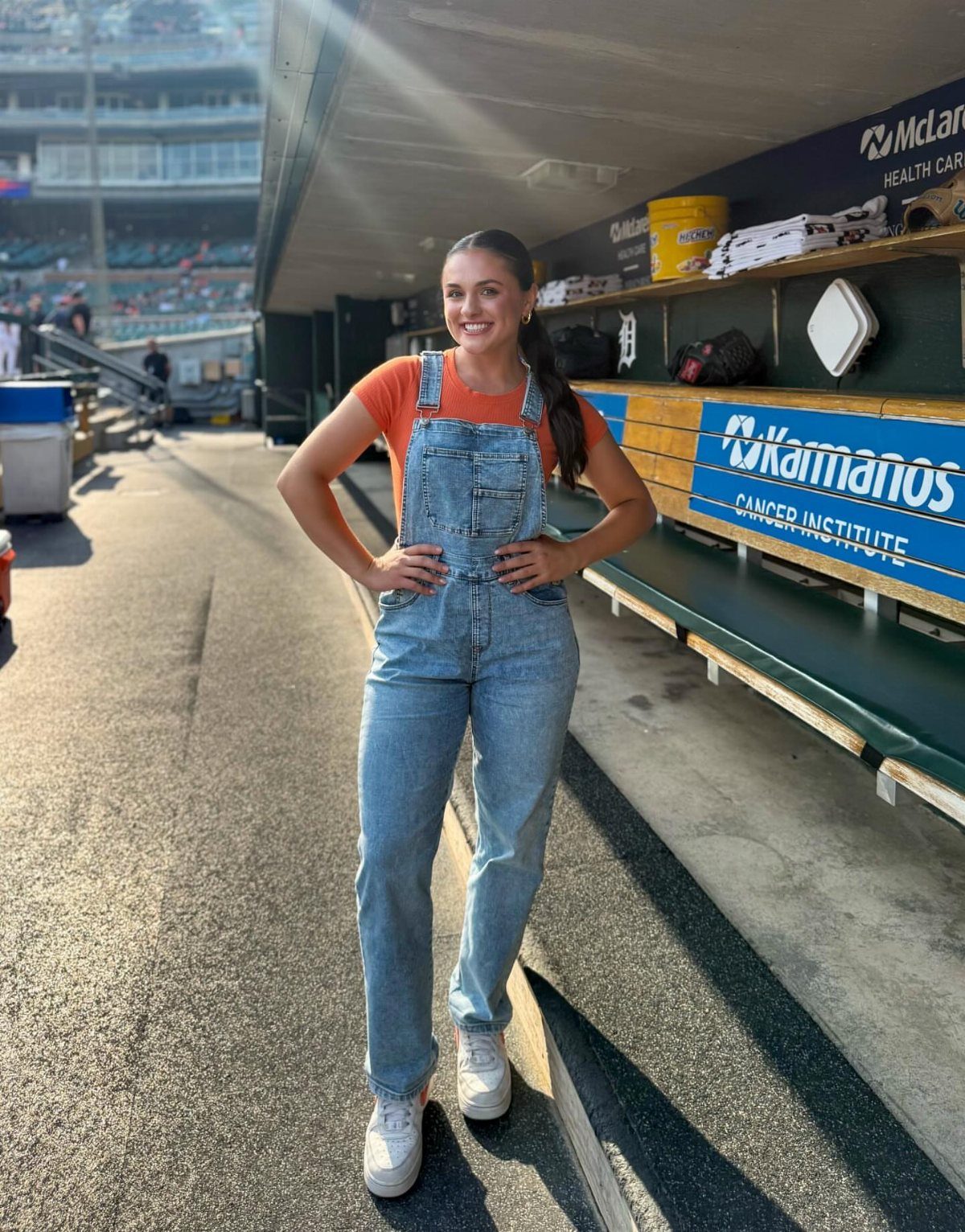 Woman in denim overalls and orange t-shirt posing in a baseball dugout.