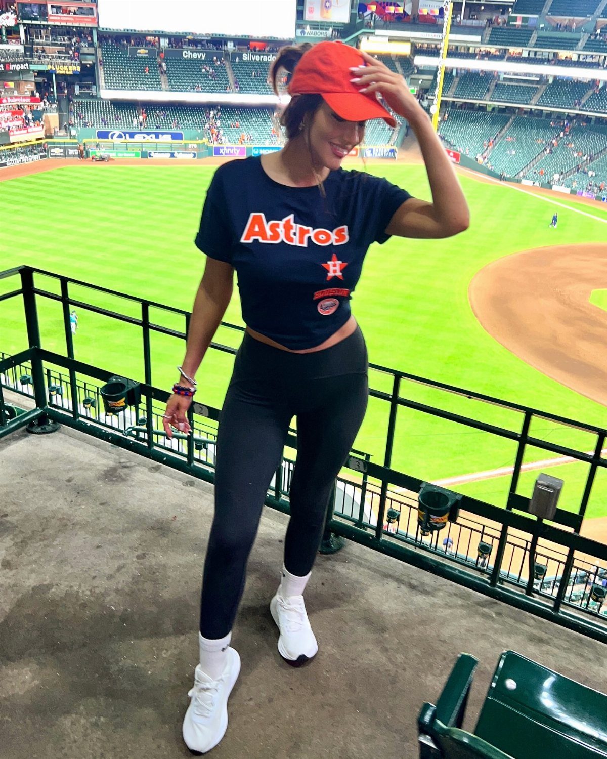 Woman in Astros t-shirt, black leggings, and red cap at a baseball stadium.