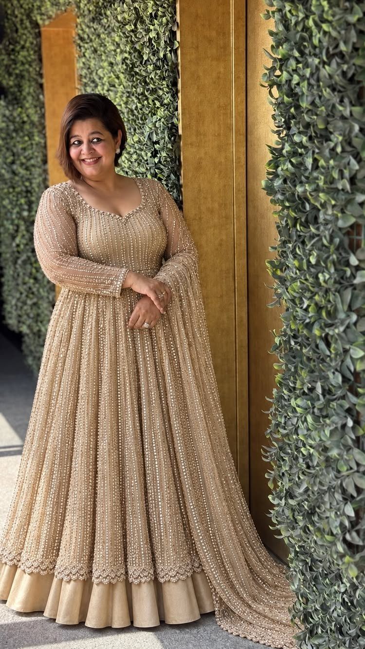 Woman in a gold beaded long-sleeved gown standing by a green hedge.