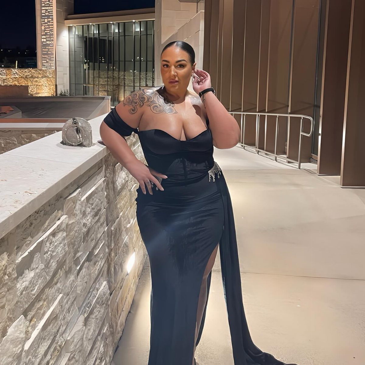 Woman in a long black off-the-shoulder gown posing by a stone wall outdoors.