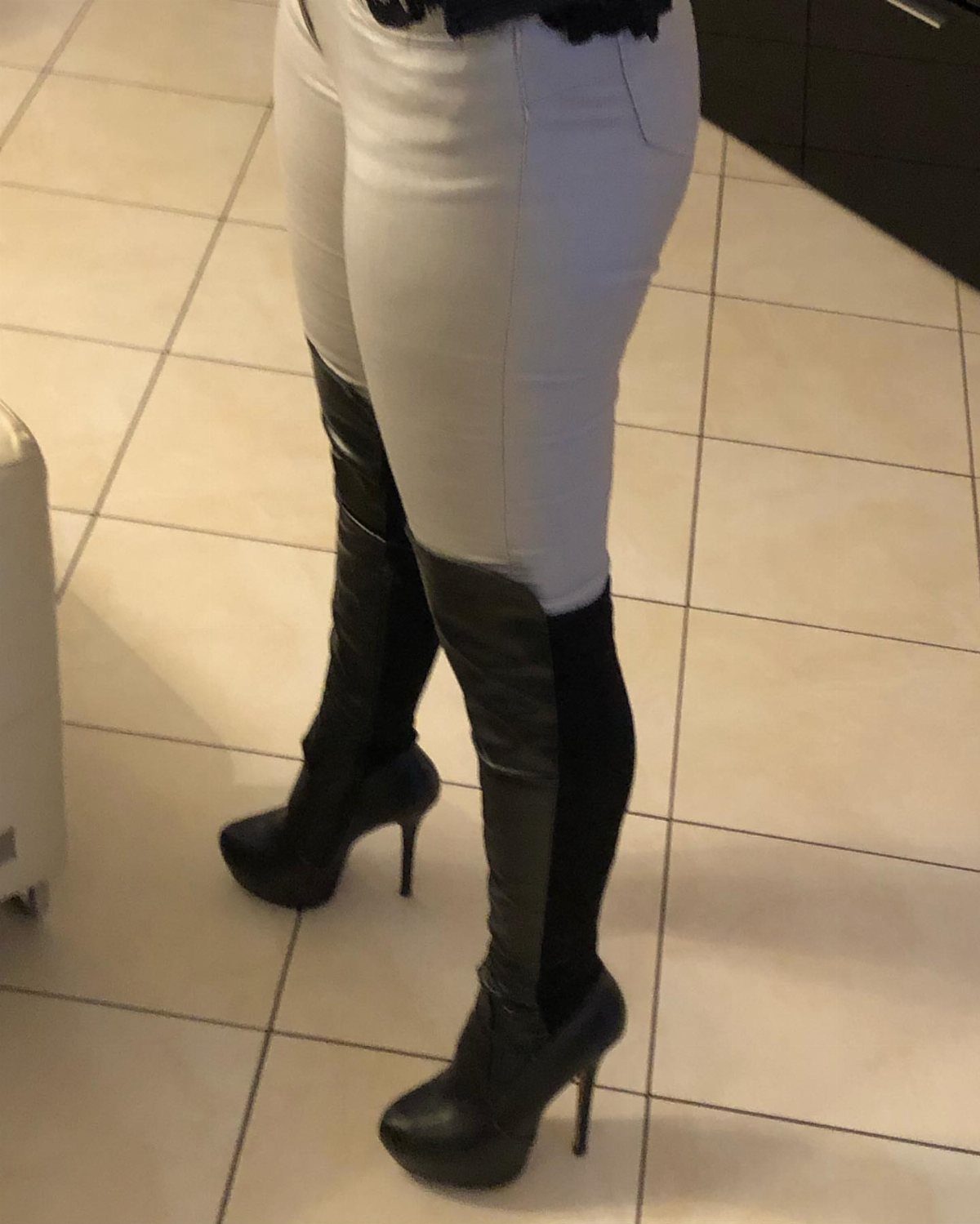 Black high-heeled platform boots worn with tight white pants on tiled flooring.
