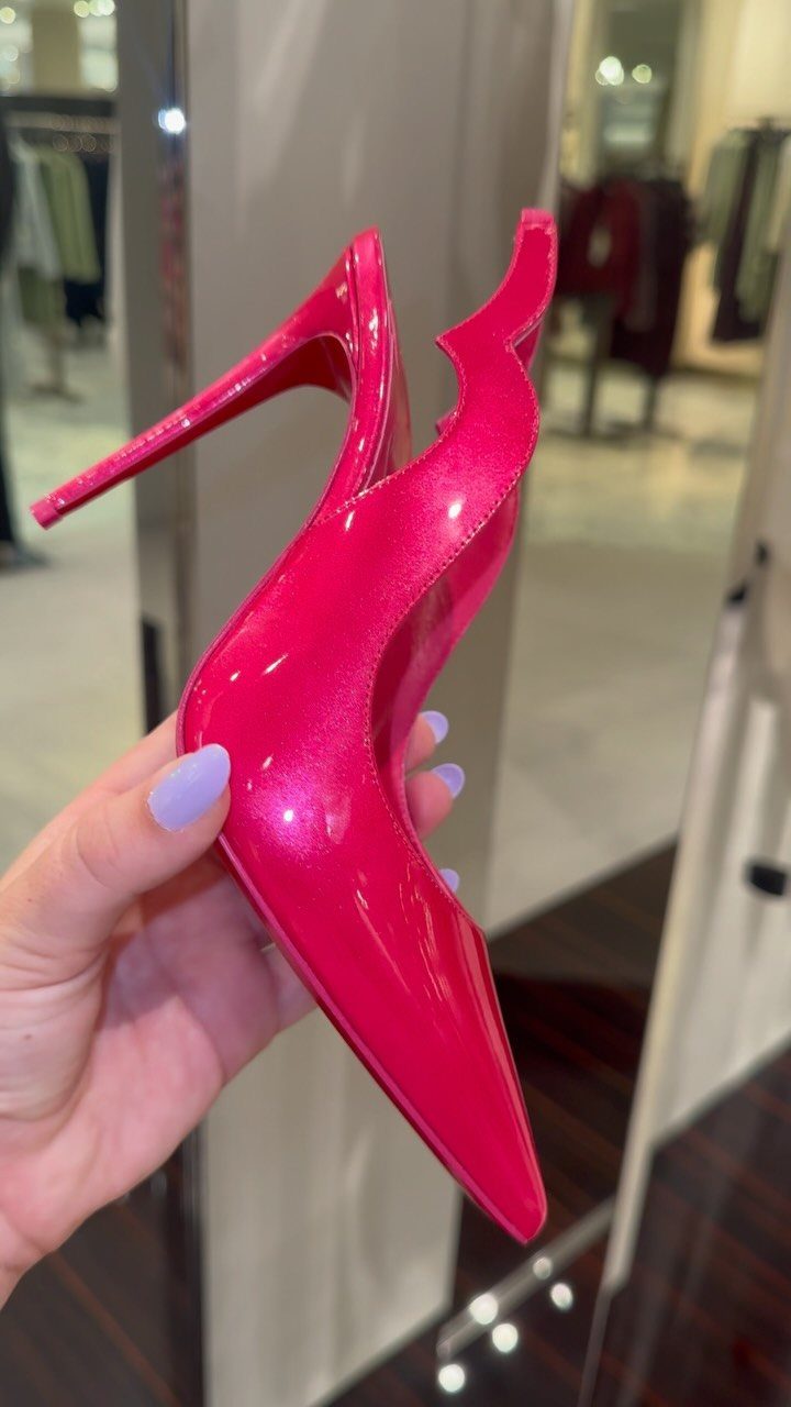 Bright pink glossy pointed-toe high heel held by a hand with purple nails.