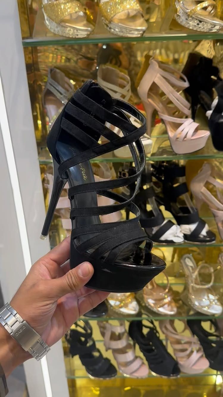 Black high-heeled sandal with criss-cross straps held in front of a shoe display.