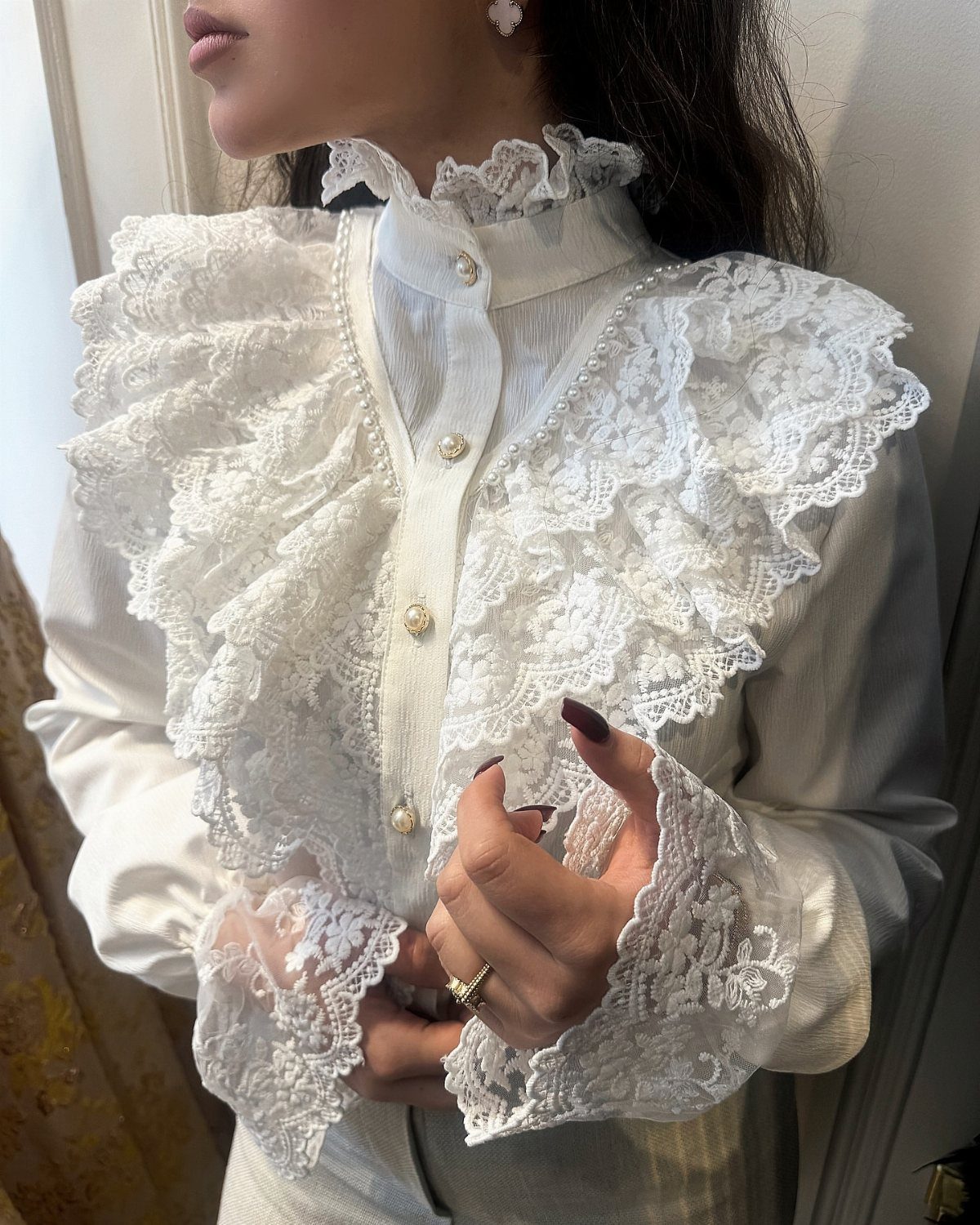 Woman wearing a white high-collared blouse with intricate lace ruffles and gold buttons.