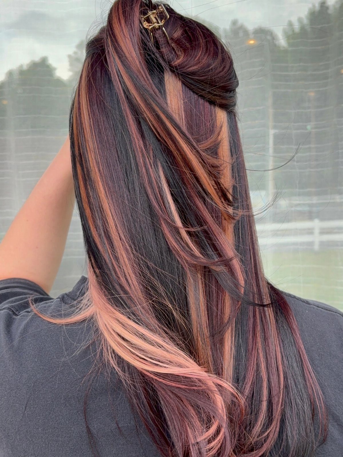 Long dark brown hair with thick vertical copper and rose-gold highlights from behind.
