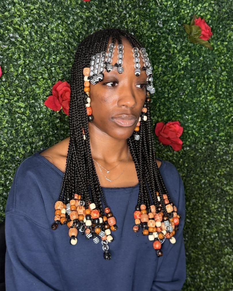 Woman with beaded cornrows and braided bangs standing before a green floral wall