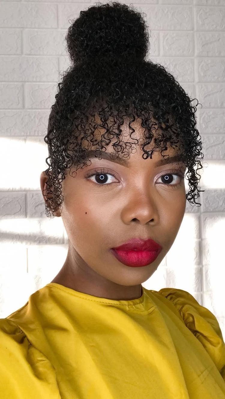 Woman with a high bun and tight curly bangs wearing a yellow top.