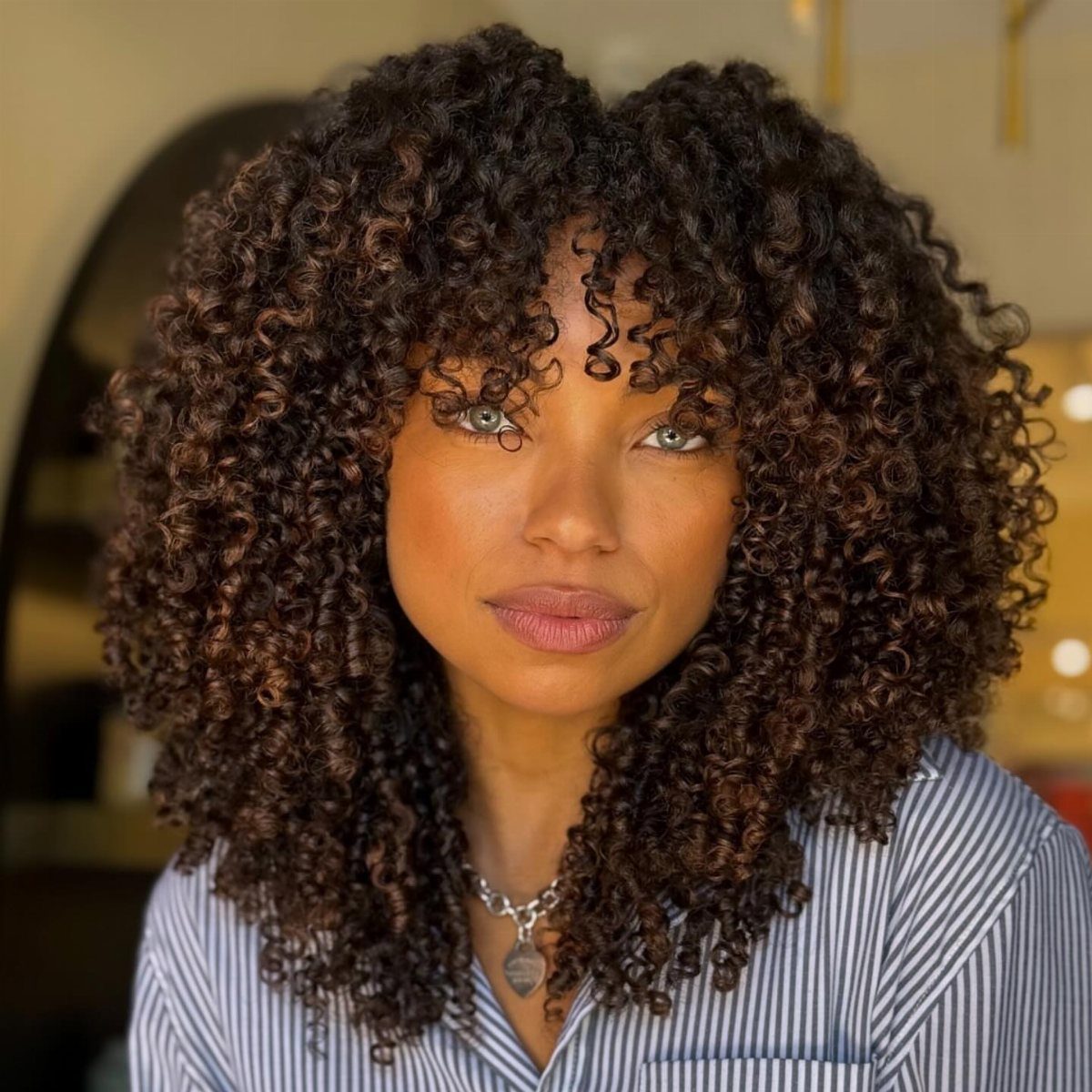Woman with voluminous dark brown curly layered hair and curly bangs posing indoors.