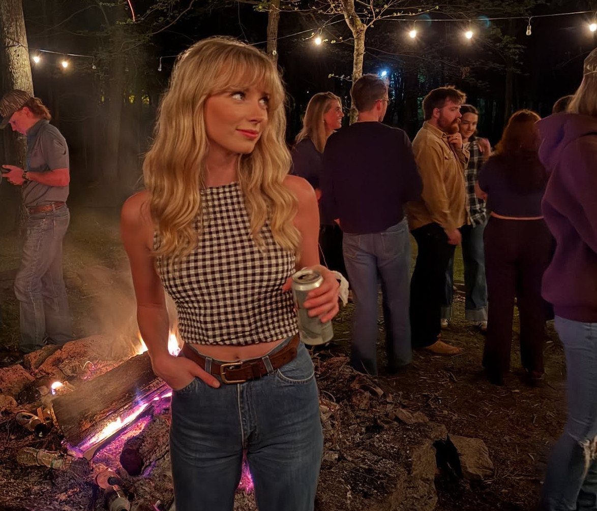 Cute Bonfire Outfits: 11 Ideas for Cozy Fall Nights 3 Blonde woman in checkered crop top and jeans standing by a night bonfire.
