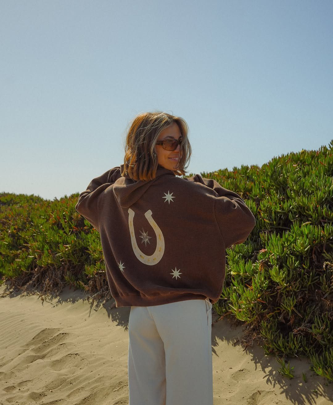 Cute Bonfire Outfits: 11 Ideas for Cozy Fall Nights 1 Woman in brown oversized hoodie with horseshoe design standing on a sandy beach