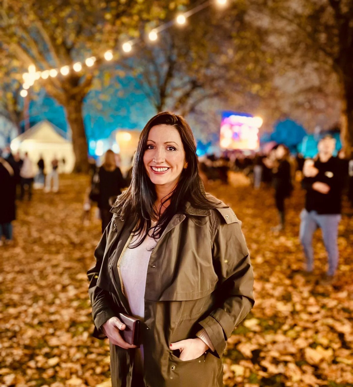 Cute Bonfire Outfits: 11 Ideas for Cozy Fall Nights 6 Woman in olive green jacket smiling in an autumn park with fallen leaves.