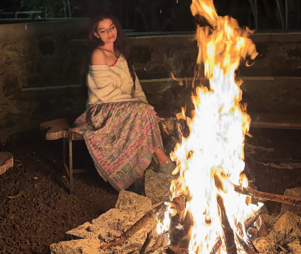 Cute Bonfire Outfits: 11 Ideas for Cozy Fall Nights 9 Woman in white sweater and patterned skirt sitting by a large outdoor bonfire