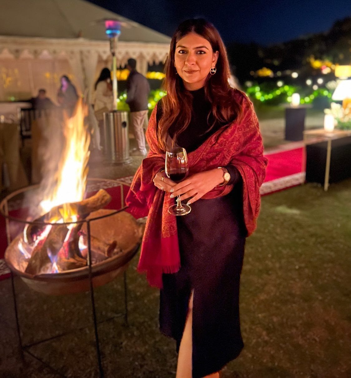 Cute Bonfire Outfits: 11 Ideas for Cozy Fall Nights 8 Woman in black dress and red shawl standing by a glowing bonfire pit.