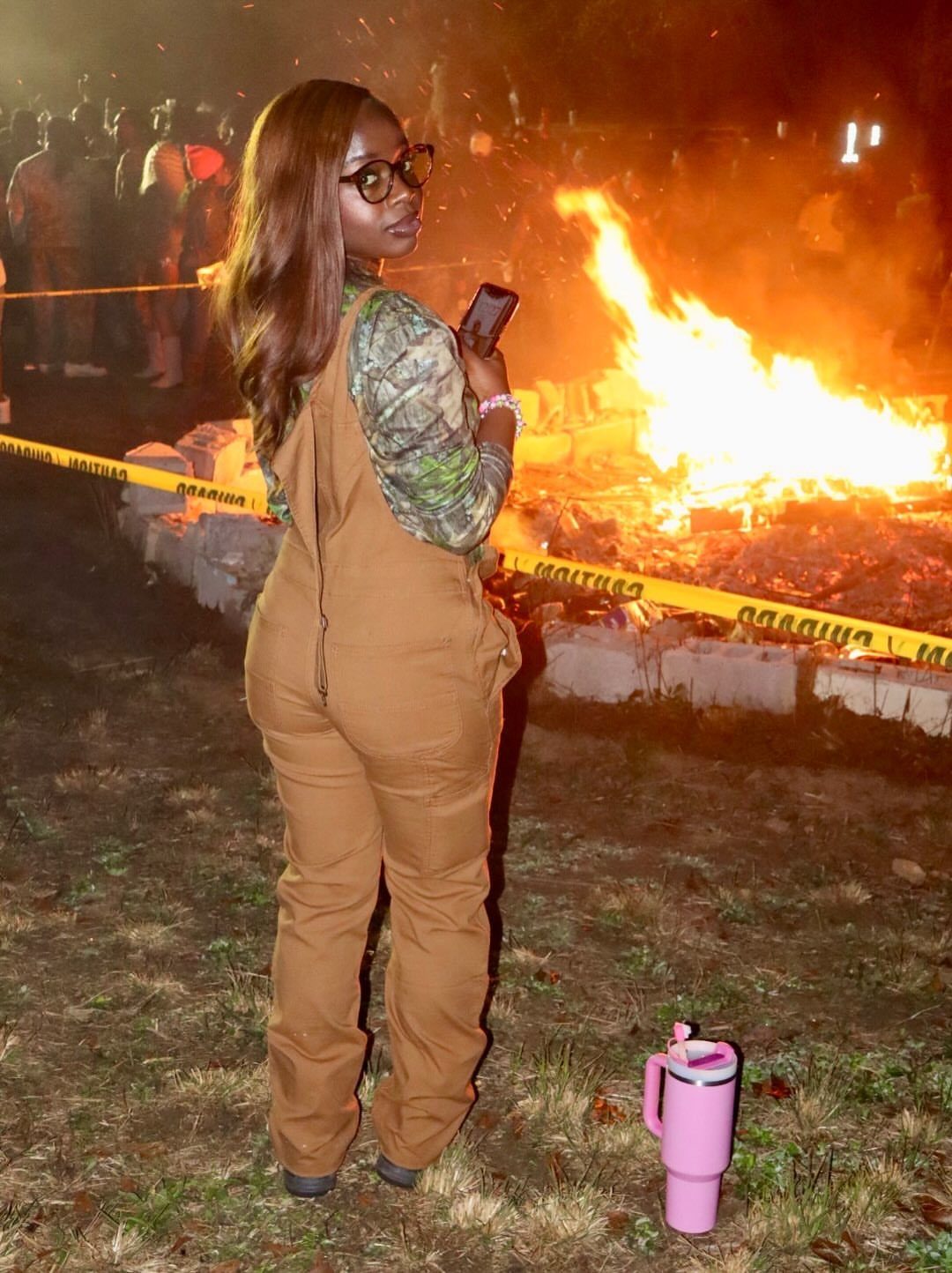 Cute Bonfire Outfits: 11 Ideas for Cozy Fall Nights 7 Woman in brown overalls standing near a large bonfire at night with pink tumbler.