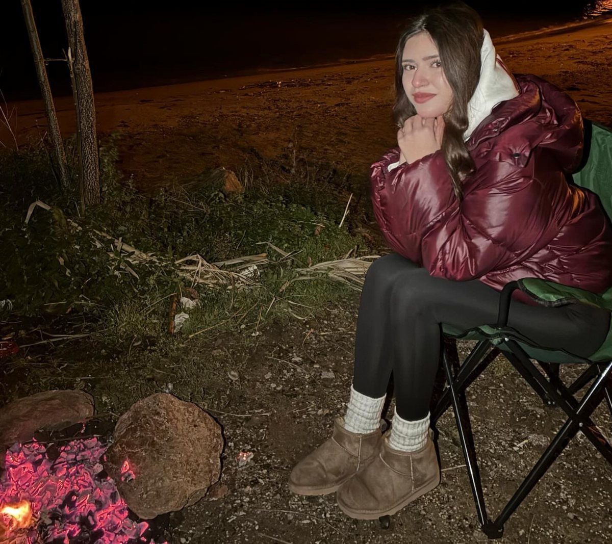 Cute Bonfire Outfits: 11 Ideas for Cozy Fall Nights 2 Woman in a burgundy oversized puffer coat sitting by a bonfire at night.