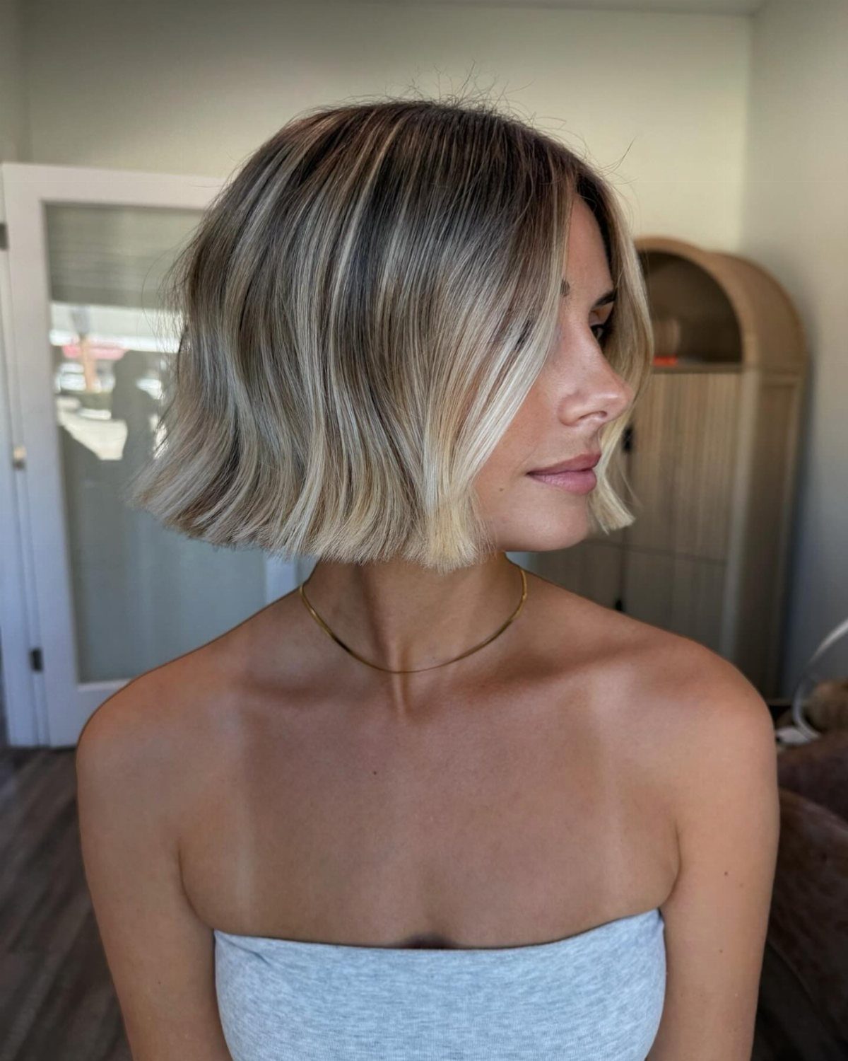 Woman with a blonde balayage A-line bob haircut and a grey strapless top.
