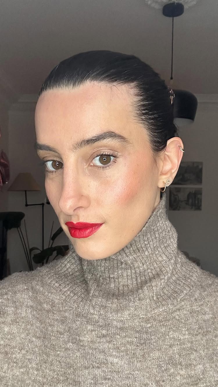 Woman with sleek wet-look dark hair, red lipstick, and a beige turtleneck sweater.