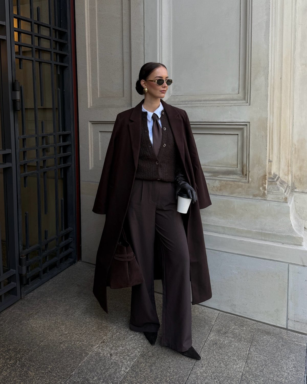 Woman in a long brown wool coat and matching wide-leg trousers standing outdoors.