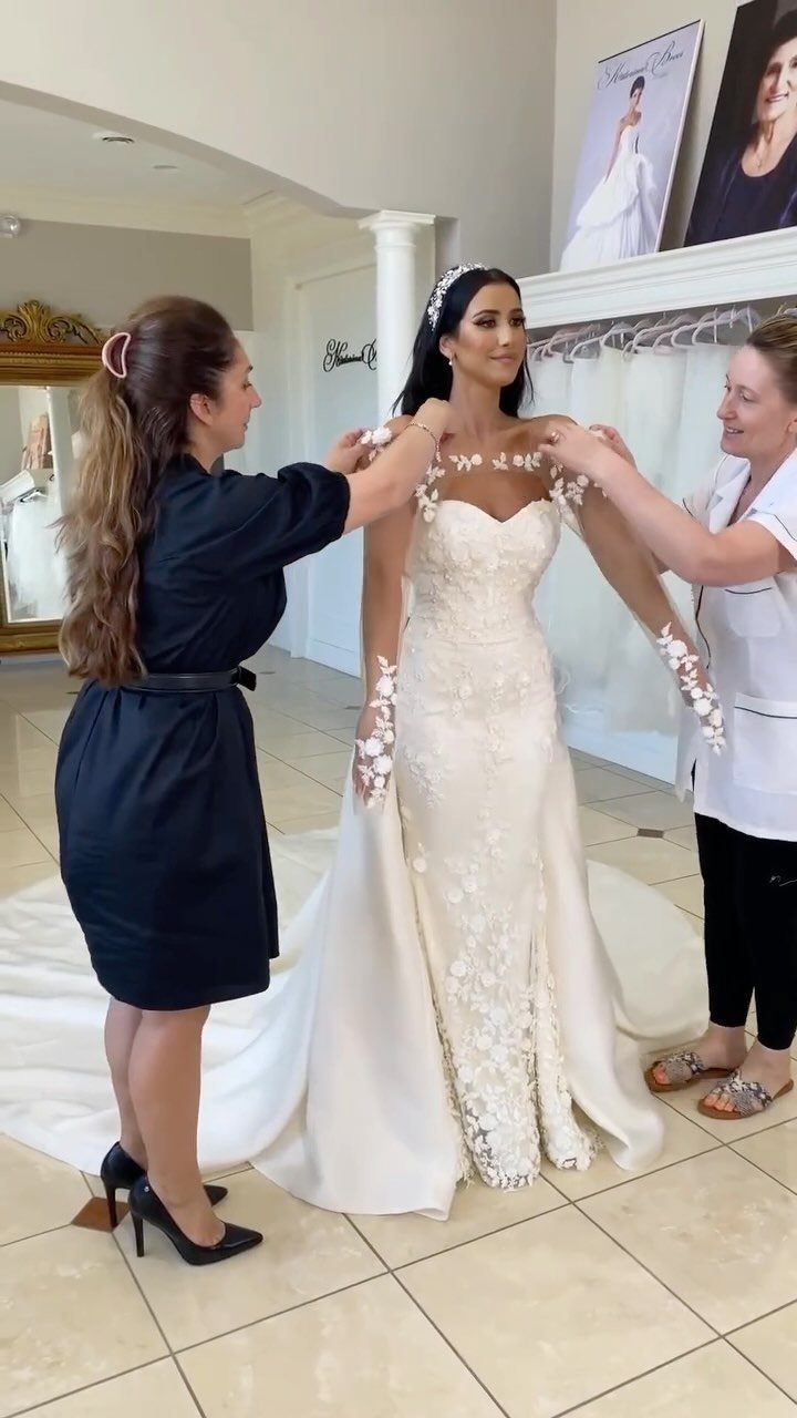 Bridal consultants fitting floral lace detachable sleeves on a white wedding gown.