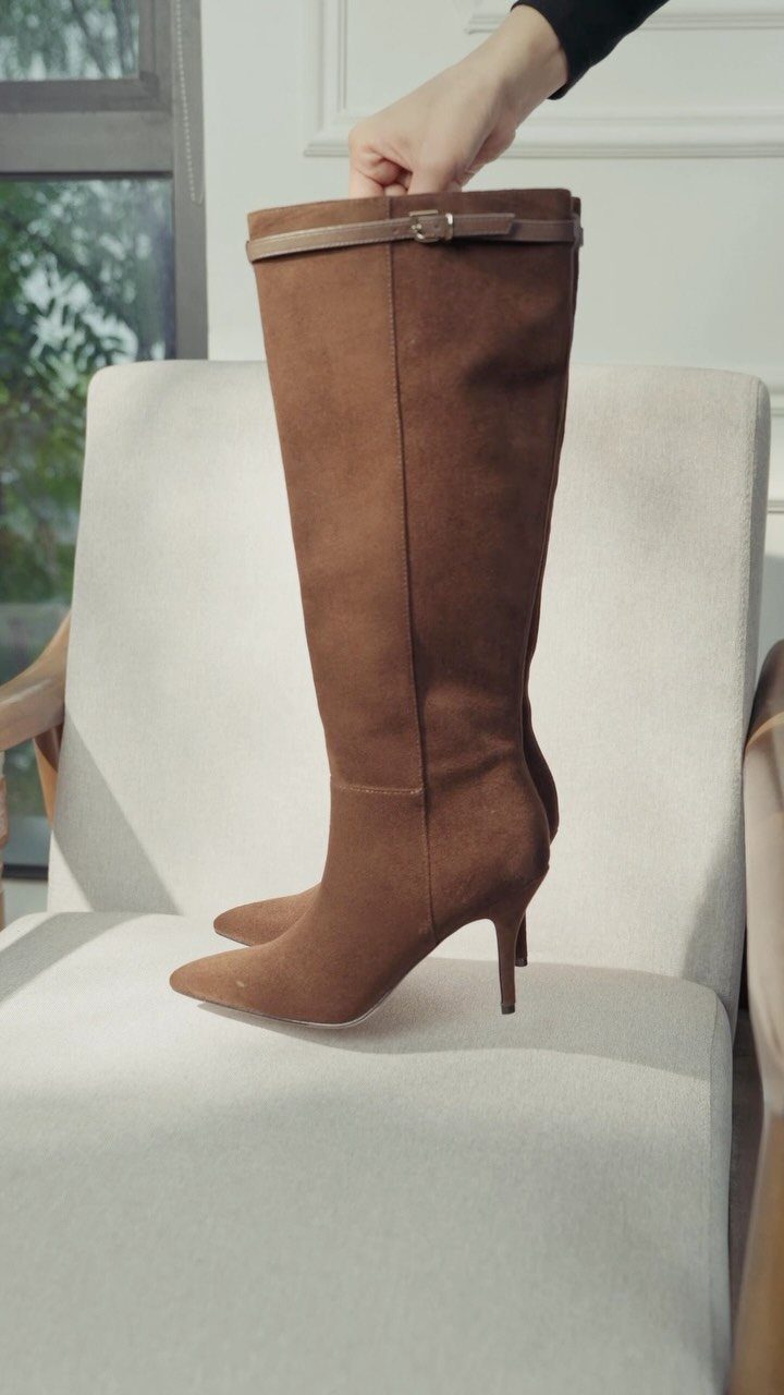 Brown suede knee-high boots with stiletto heels resting on a beige armchair.