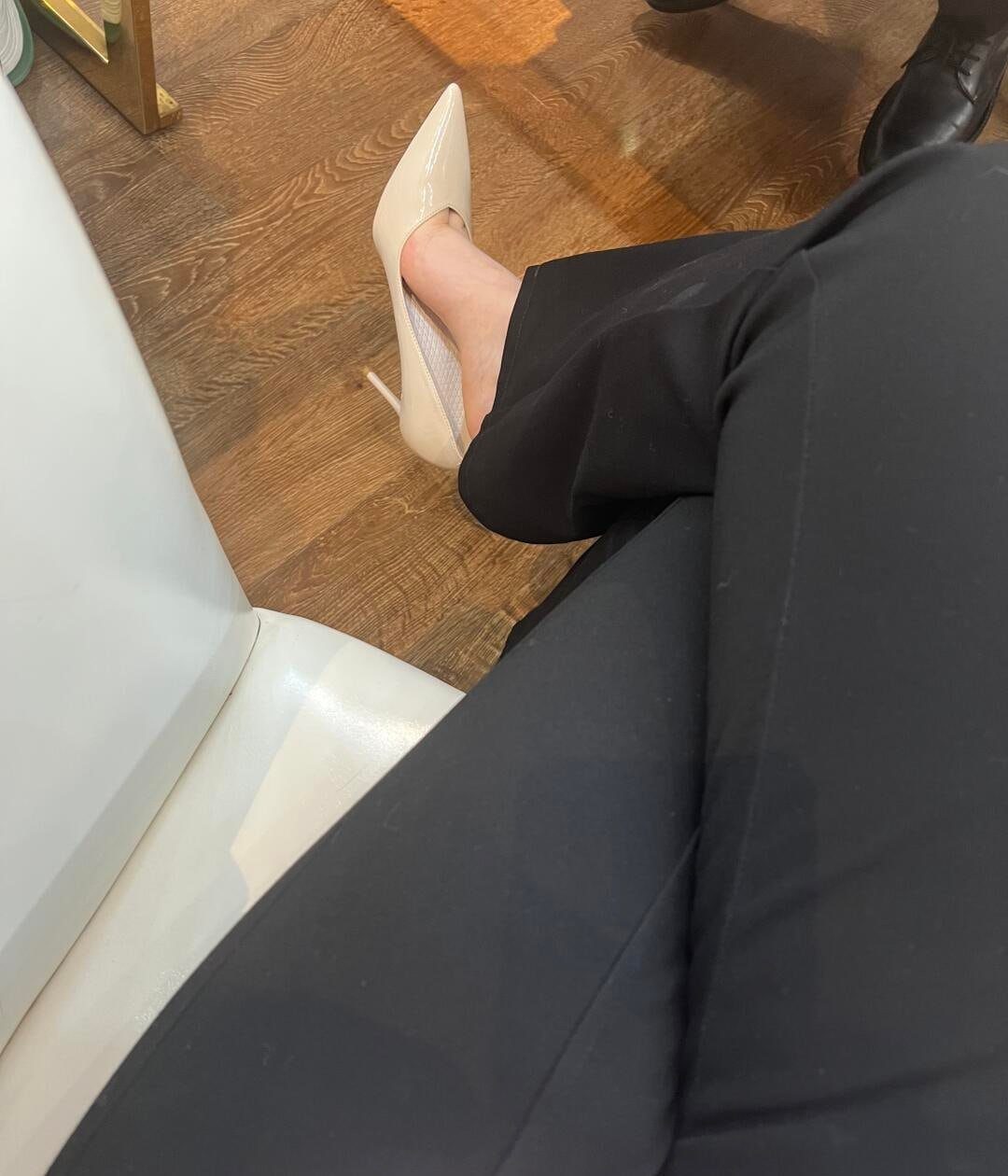 Beige pointed-toe high heel shoe worn with black trousers on wooden flooring.