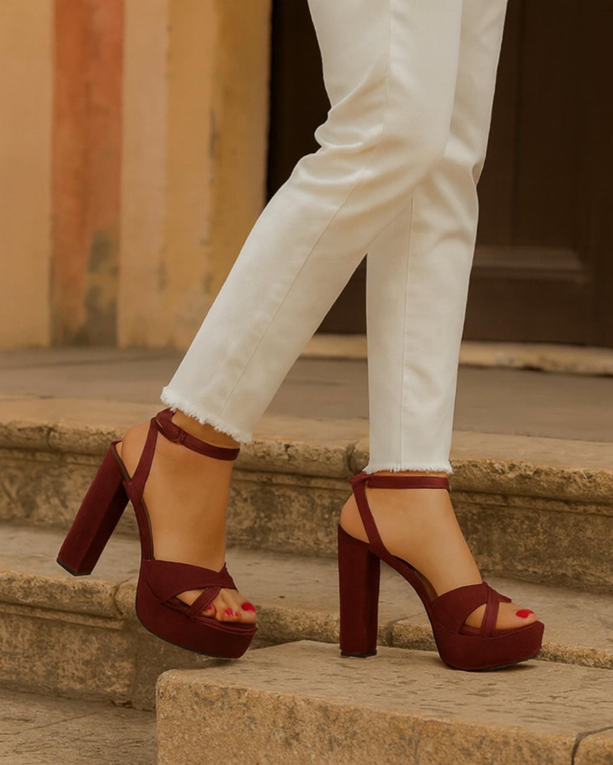 Burgundy suede platform sandal heels with ankle straps worn with white trousers.