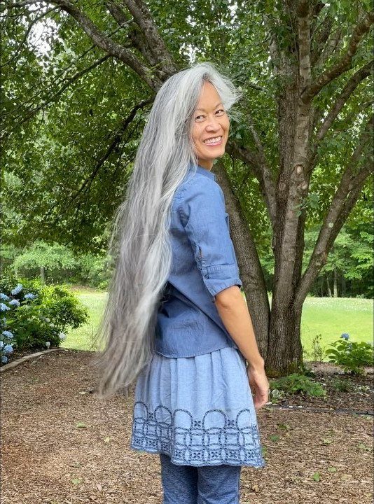 Woman with very long grey hair smiling outdoors in a blue denim outfit.