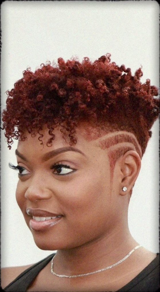 Woman with curly maroon hair and shaved side designs posing against light background