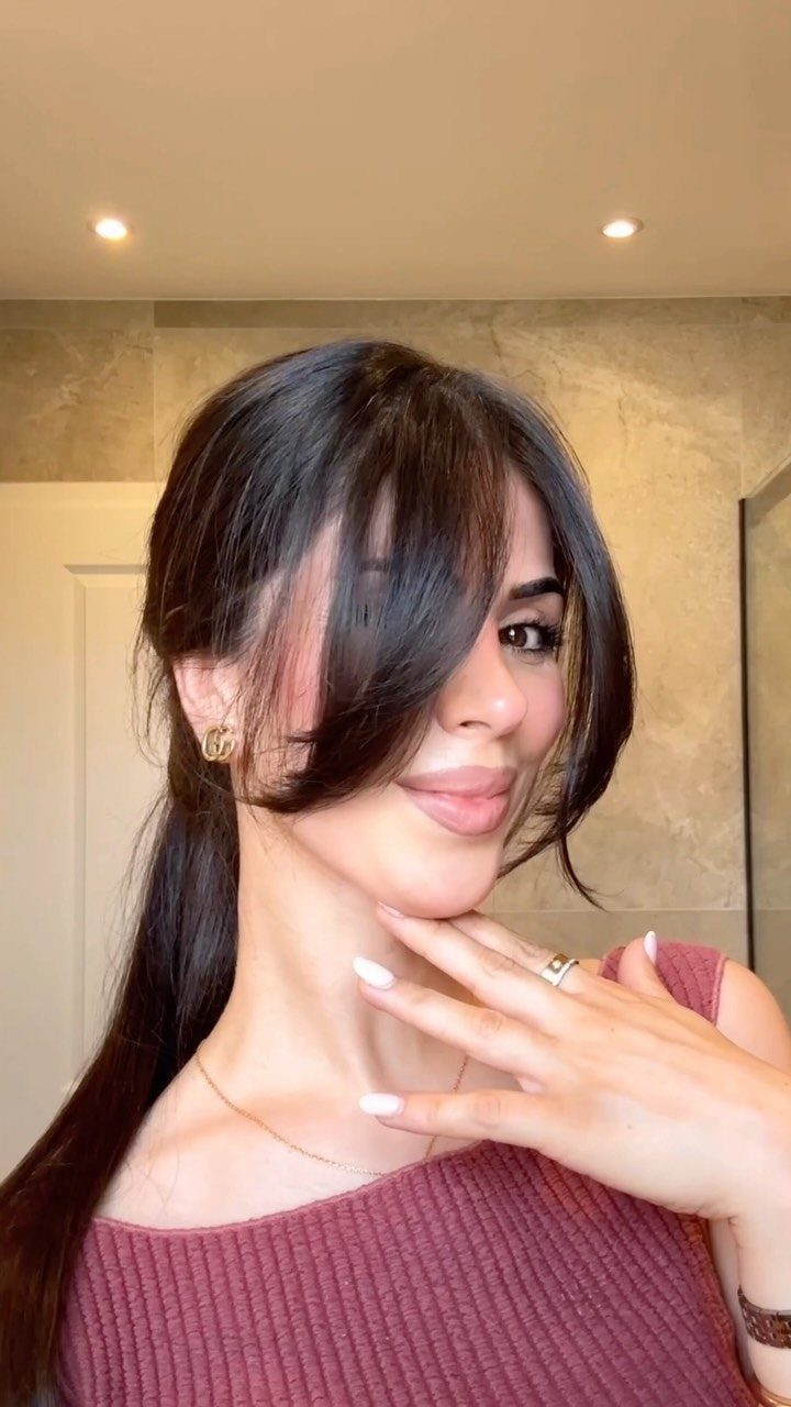 Woman with dark hair in a ponytail and long face-framing bangs wearing mauve top.
