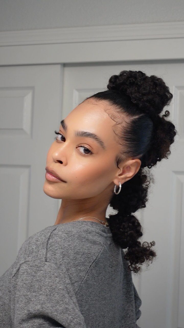 Woman with dark curly hair in a high slicked-back ponytail wearing grey sweater