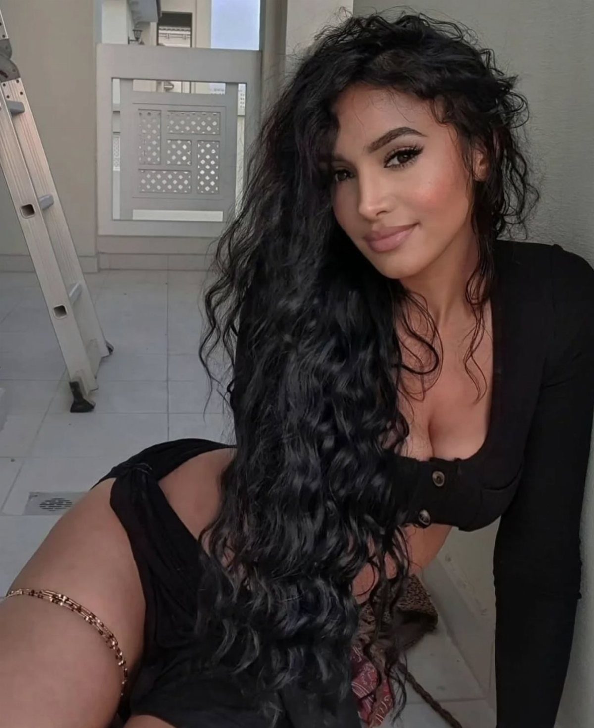 Woman with long, thick, dark curly hair wearing a black top and gold jewelry.