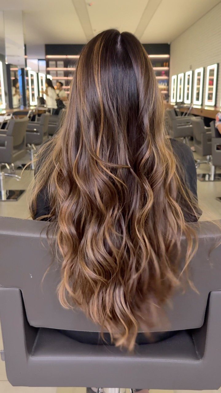 Long wavy brown hair with hazelnut balayage highlights in a hair salon setting.