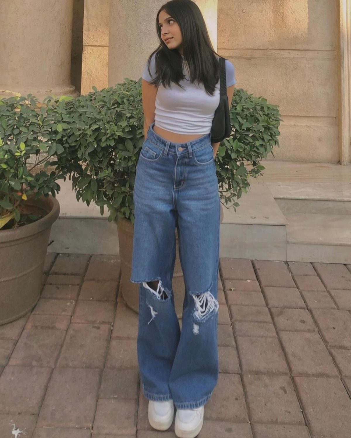 Woman wearing high-waisted ripped blue jeans, a white cropped tee, and white sneakers.
