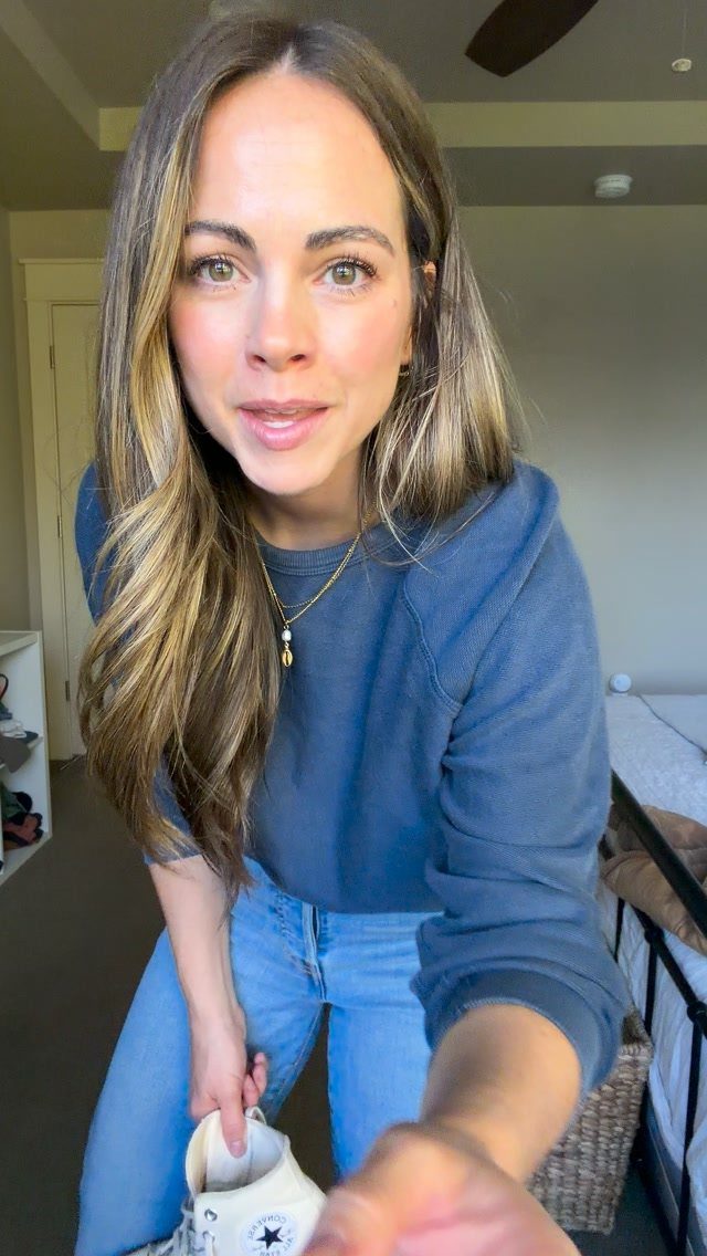 Woman wearing a blue sweatshirt and light blue jeans holding a white sneaker.
